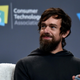 Jack Dorsey sitting in front of a wall that says CES staring off and smiling with a microphone in his hand.